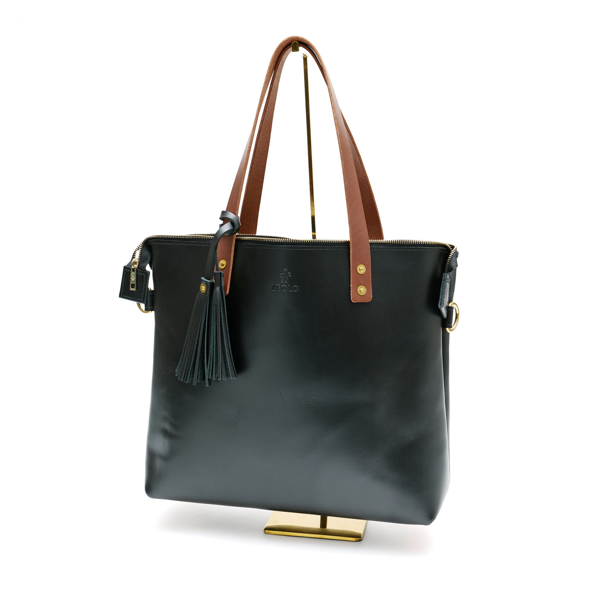 Black leather tote bag with brown handle and black tassel charm, on bag stand. 