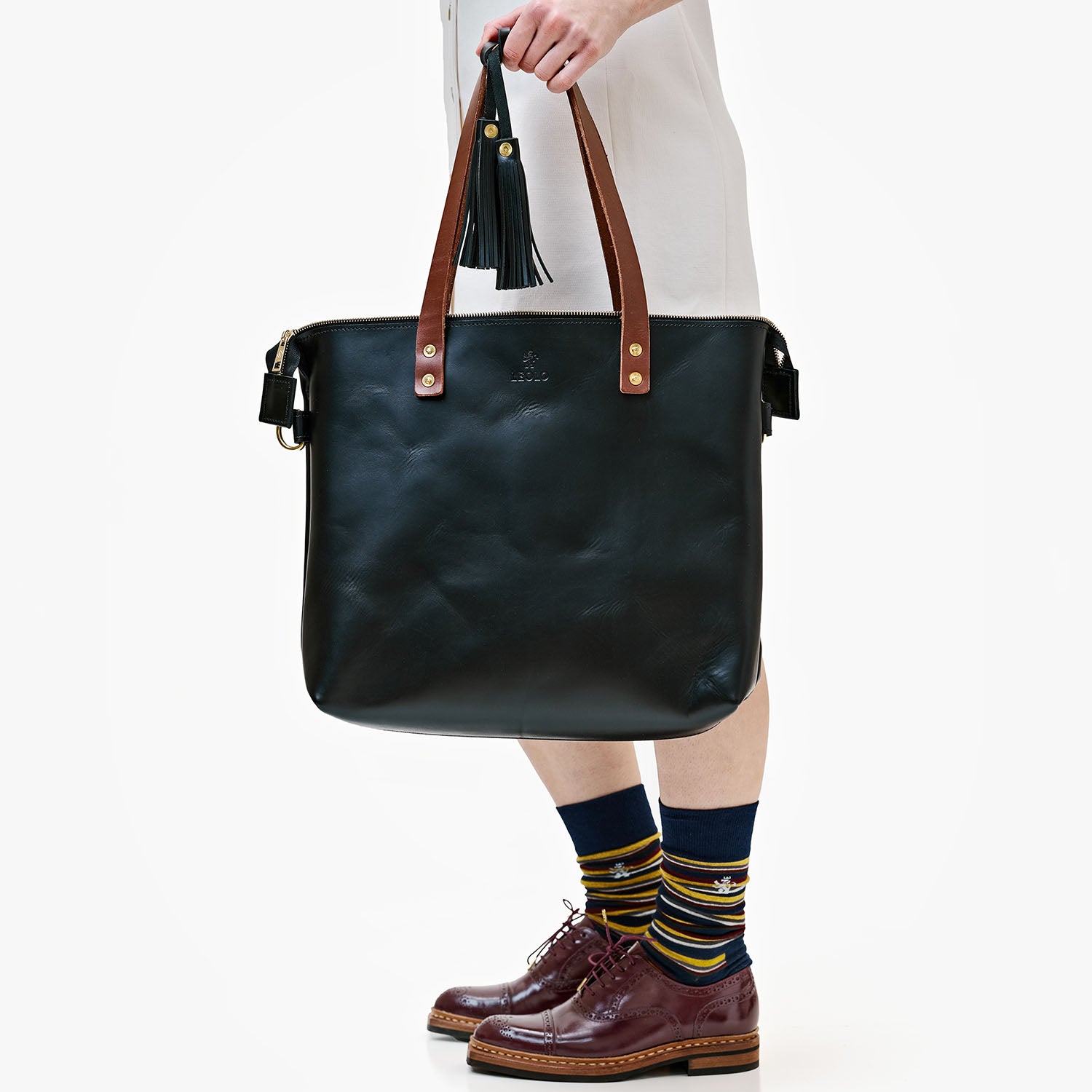 Person in front of white background holding black leather tote bag with brown straps.