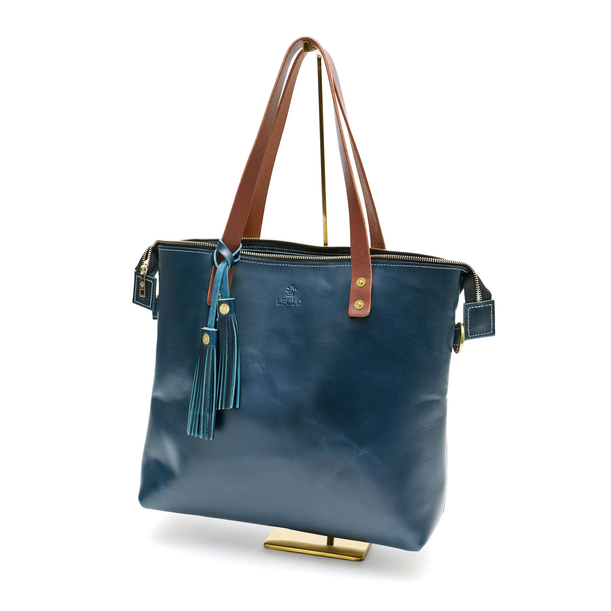 Deep blue leather tote bag with brown handle and blue tassel charm, on bag stand. 