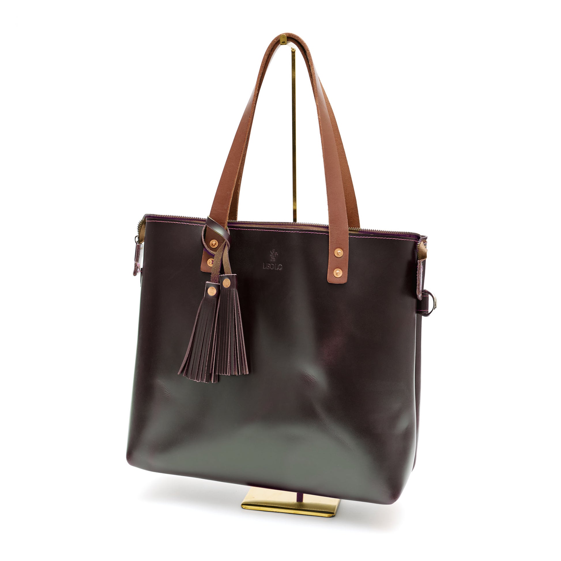 Brown leather tote bag with brown handle and tassel charm, on bag stand. 