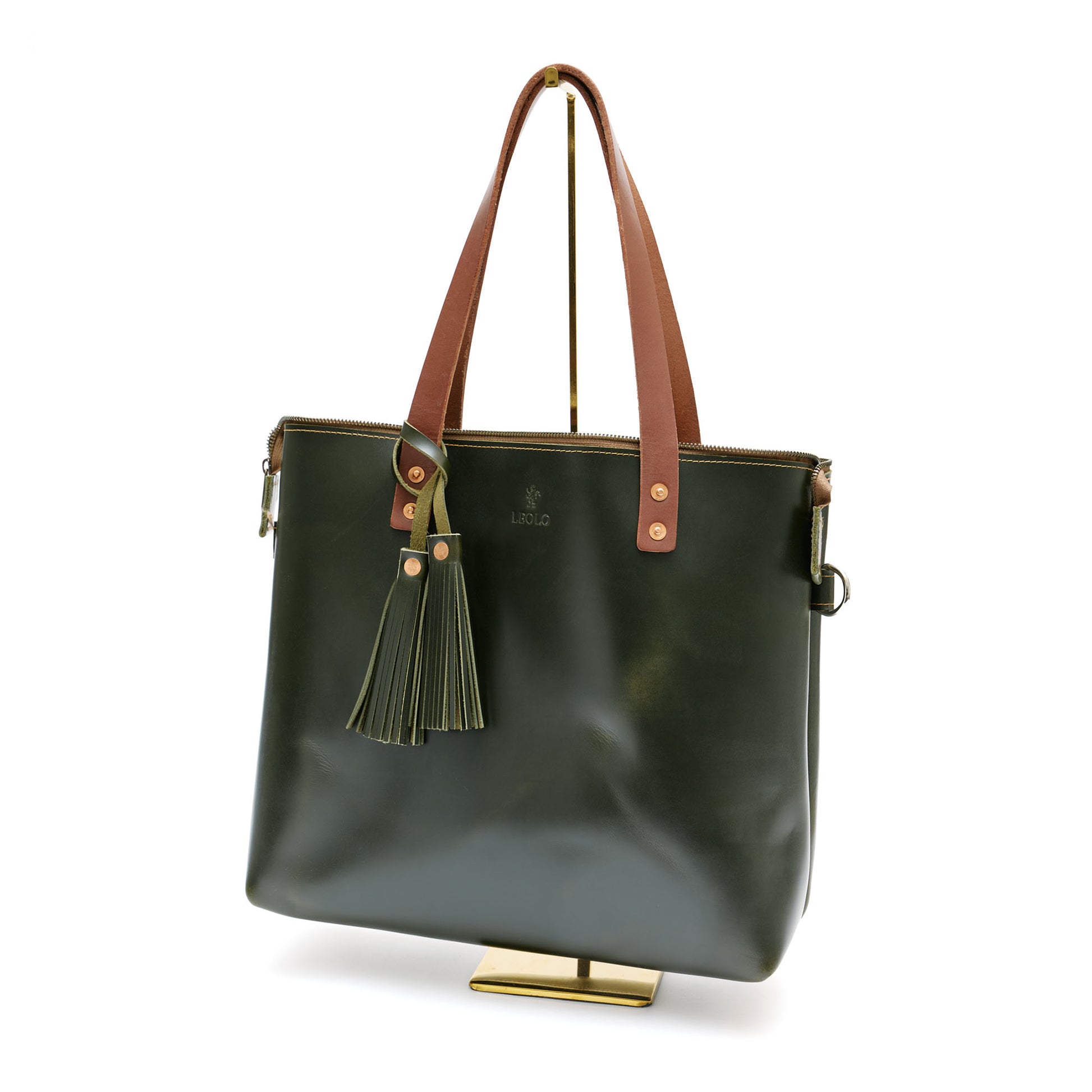 Olive green leather tote bag with brown handle and green tassel charm, on bag stand. 